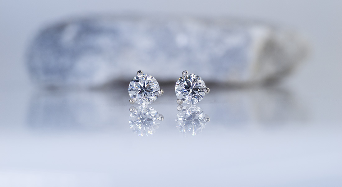 A pair of diamond stud earrings in 14k white gold martini settings in front of a rock on a light background